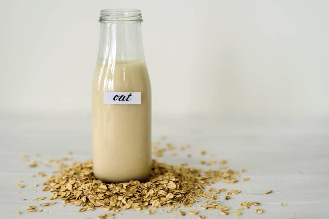 Glass bottle of oat milk on white background behing heap of oatmeal flakes