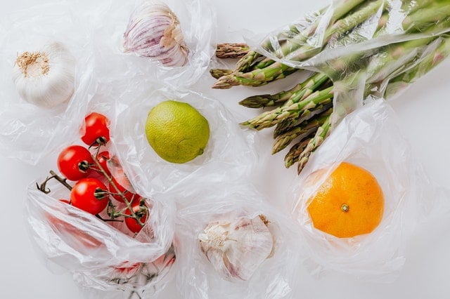 cherry tomatoes, garlic, asparagus, and an orange in plastic bags on white surface