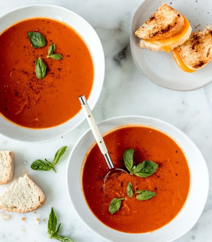 Two white bowls filled with healthy tomato soup, with a grilled cheese on a plate next to them. 